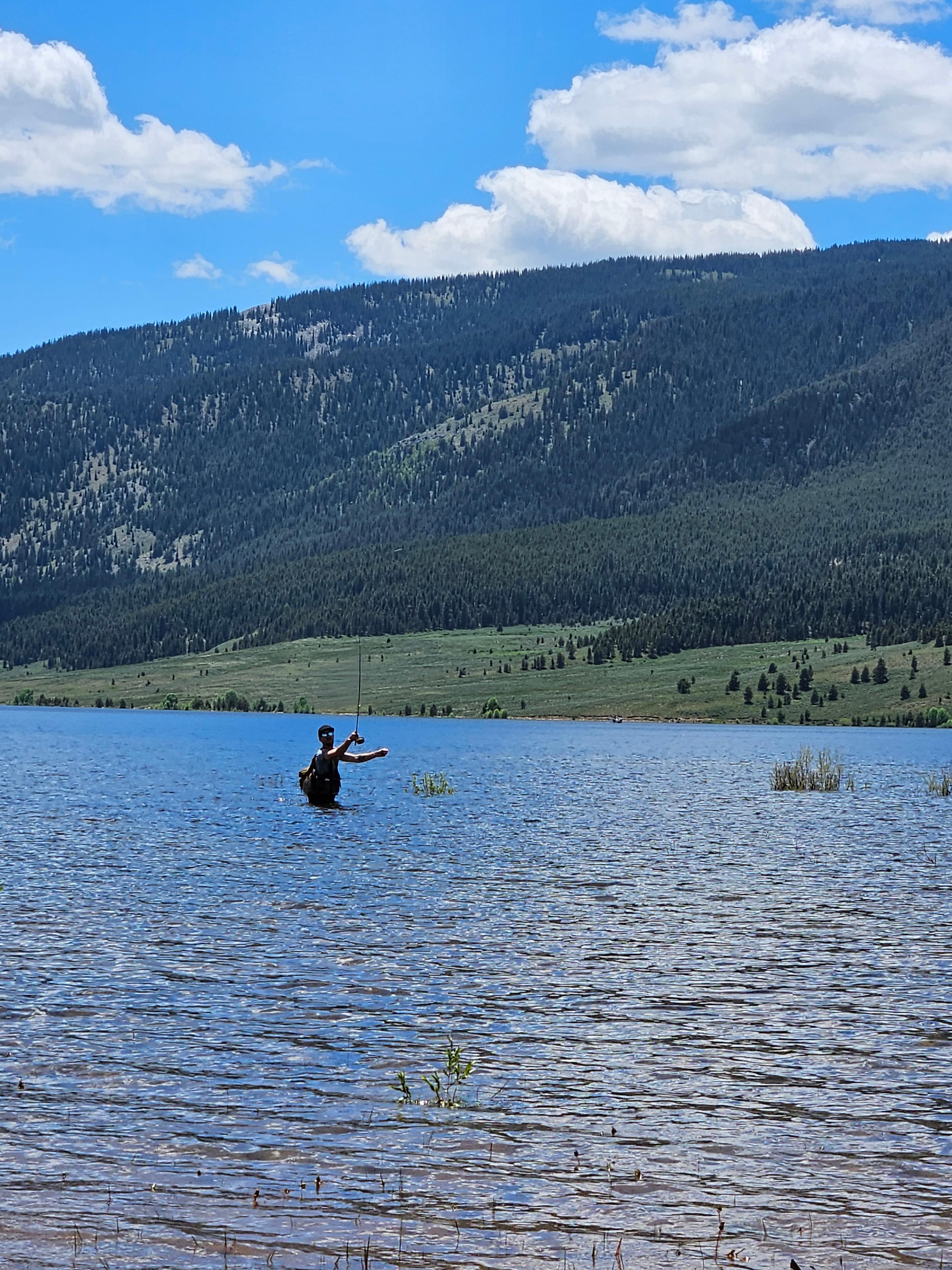 Angler fly fishing in a serene lake, casting towards the shoreline under a setting sun, using strategic techniques for stillwater success.