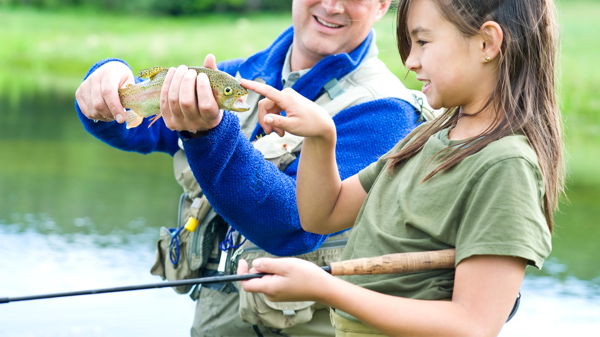 a father holding a rainbow trout letting his daughter see the trouts spots up clost. She is holding a fly rod. 