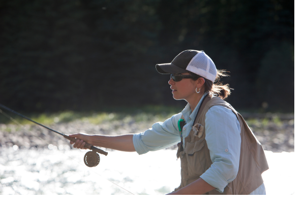 An angler casting a fly rod into a clear mountain stream, with a backdrop of rocky hills and lush greenery along the banks.